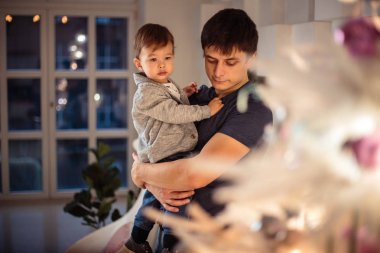 Küçük oğlu rahat bir ceket ve kot babasıyla birlikte Noel ağacının yanında oturuyor. Genç adam ve oğlu