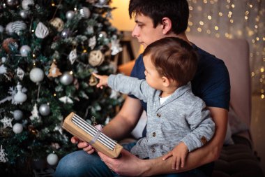 Küçük oğlu rahat bir ceket ve kot babasıyla birlikte Noel ağacının yanında oturuyor. Genç adam ve oğlu