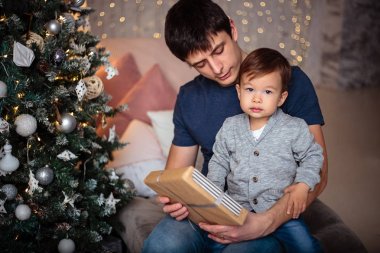 Küçük oğlu rahat bir ceket ve kot babasıyla birlikte Noel ağacının yanında oturuyor. Genç adam ve oğlu