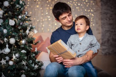 Küçük oğlu rahat bir ceket ve kot babasıyla birlikte Noel ağacının yanında oturuyor. Genç adam ve oğlu