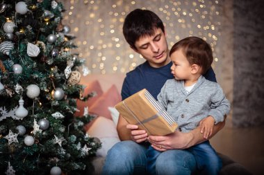Küçük oğlu rahat bir ceket ve kot babasıyla birlikte Noel ağacının yanında oturuyor. Genç adam ve oğlu