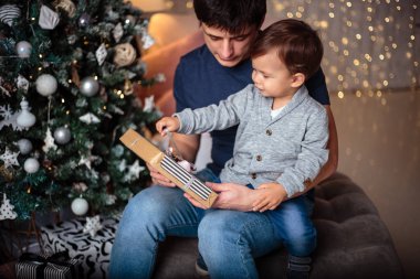 Küçük oğlu rahat bir ceket ve kot babasıyla birlikte Noel ağacının yanında oturuyor. Genç adam ve oğlu