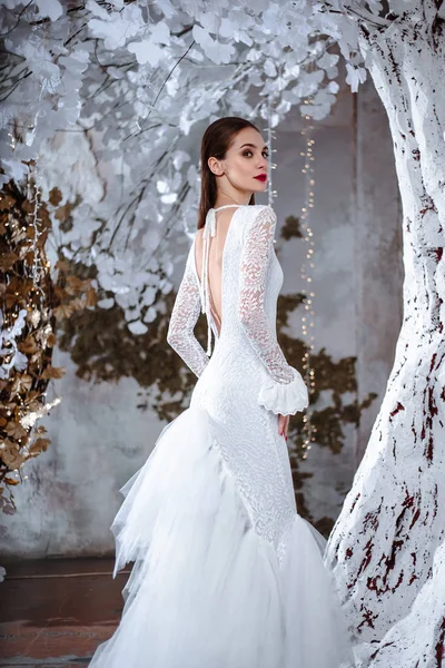 Fashion portrait of beautiful young woman in a gorgeous wedding dress, posing in a fantasy interior with white winter tree