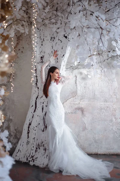 Fashion portrait of beautiful young woman in a gorgeous wedding dress, posing in a fantasy interior with white winter tree