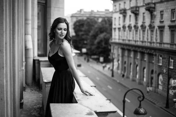 Black white portrait of young woman in elegant silk black dress lookingto a city from a balcony, fashion beauty photo