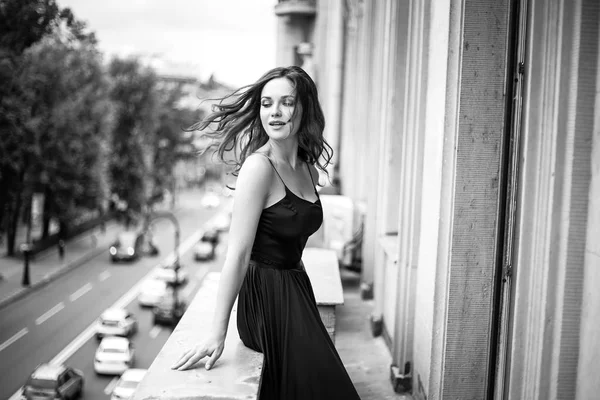 Black white portrait of young woman in elegant silk black dress lookingto a city from a balcony, fashion beauty photo