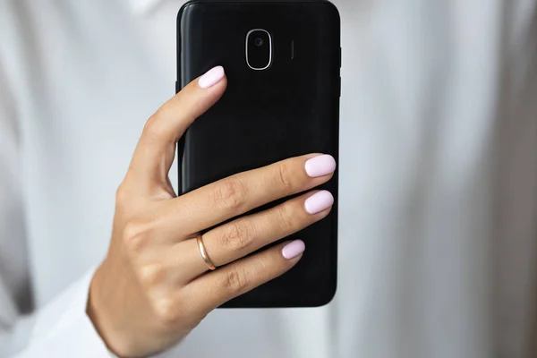 Close-up photo of elegant light pink manicure over white shirt background, tender women\'s hands with perfect nails hold a mobile phone