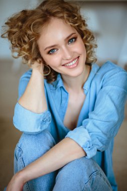 Kıvırcık saçlı ve büyüleyici gülümseme ile Oldukça neşeli sarışın genç kadın evinde dinleniyor, modern İskandinav iç oda