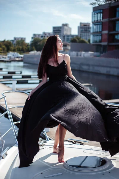 年轻的漂亮女人穿着丝质黑色的长裙 在游艇上摆姿势 奢华的生活方式 — 图库照片