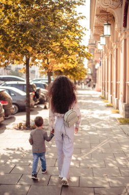 Küçük çocuk annesiyle birlikte sonbahar parkında yürüyor.