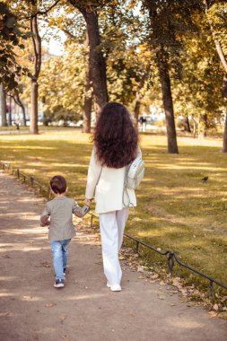 Küçük çocuk annesiyle birlikte sonbahar parkında yürüyor.