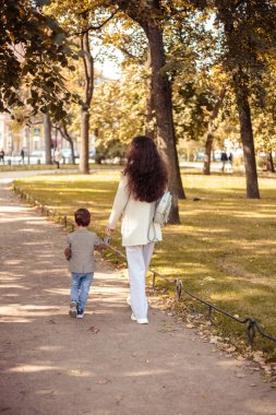 Küçük çocuk annesiyle birlikte sonbahar parkında yürüyor.