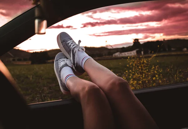 young woman with sneakers with feet propped on the car window at sunset