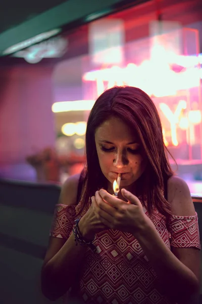 Young redhead woman lighting a cigar with a lighter next to a club with a window with neon lights