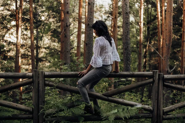 Giovane donna seduta su una recinzione di legno nella foresta tra i pini — Foto Stock