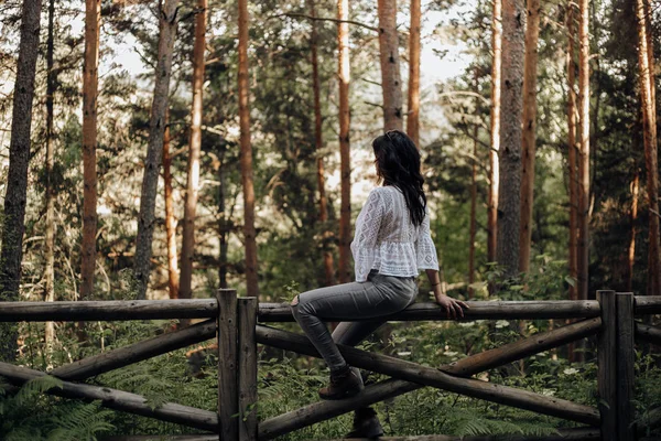 Donna seduta su una recinzione di legno nella foresta tra pini che guardano la natura — Foto Stock