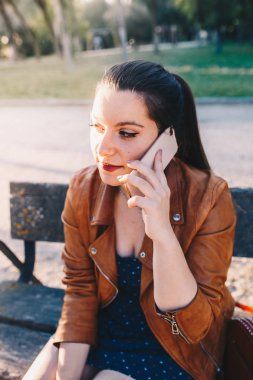Bir şehir parkında bir bankta oturan deri ceket konuşma ve akıllı telefon akıllı telefon dinleme ile Mutlu genç kadın