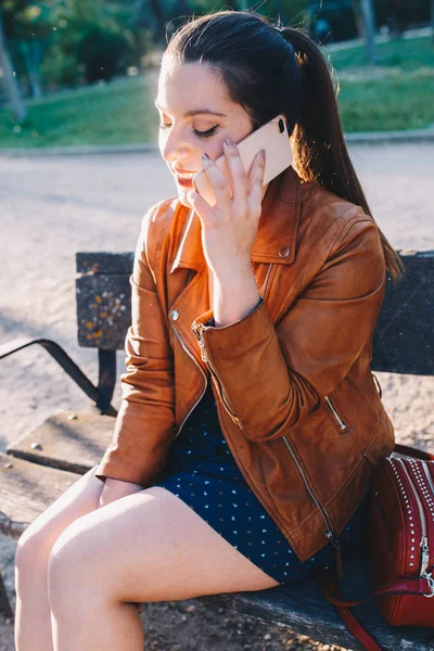 Feliz joven mujer hablando y sonriendo en el teléfono inteligente teléfono inteligente en un parque de la ciudad sentado en un banco — Foto de Stock