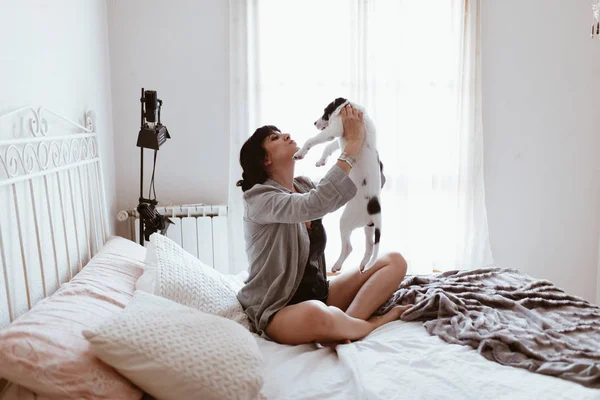 ผู้หญิงสีน้ําตาลสวย เล่นกับลูกหมา ขณะที่นั่งบนเตียงในห้องนอน — ภาพถ่ายสต็อก