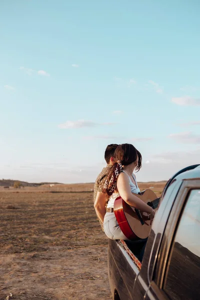 Jóvenes amantes de los viajeros que se divierten tocando la guitarra en la parte superior del coche jeep 4x4. Pareja que hace un wanderlust vacaciones en la puesta del sol en verano —  Fotos de Stock