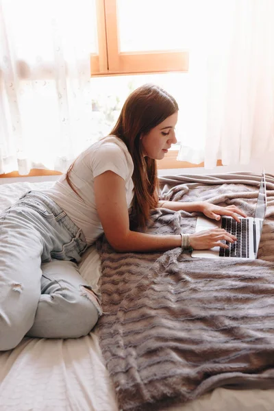 ผู้หญิงสวยนอนบนเตียงที่อบอุ่นของเธอ เขียนบนแล็ปท็อปของเธอ — ภาพถ่ายสต็อก