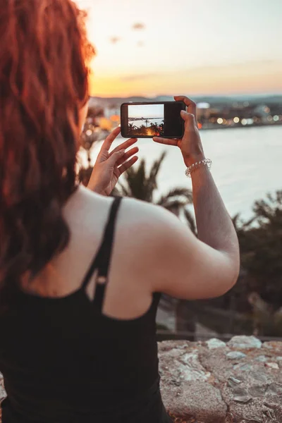 ผู้หญิงสวยถ่ายรูปภูมิทัศน์ทะเลด้วยโทรศัพท์มือถือ ในแสงที่อบอุ่นของค่ําคืนพระอาทิตย์ตก — ภาพถ่ายสต็อก