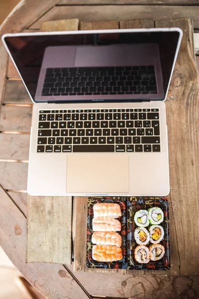 ม้วนซูชิอร่อยกุ้งและแล็ปท็อปที่โต๊ะไม้ จัดส่งอาหาร สถานที่ทํางานที่บ้าน — ภาพถ่ายสต็อก