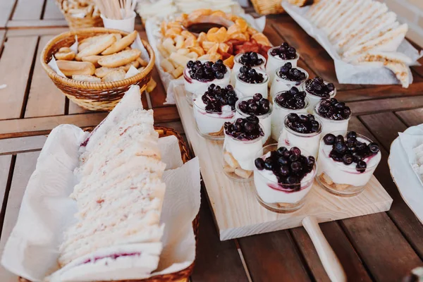Appetizers table with italian antipasti snacks. Yogurt with blueberries, variety board over rustic wooden background. Closeup of cutting board with bread, sandwiches, donuts, yogurt, fruits chocolate