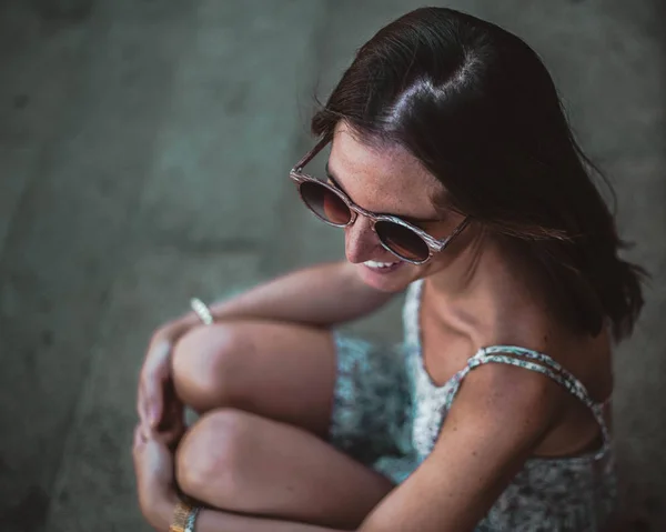 Primer plano de la hermosa chica morena en gafas de sol sentado en las escaleras. Ella mirando hacia otro lado y sonriendo — Foto de Stock