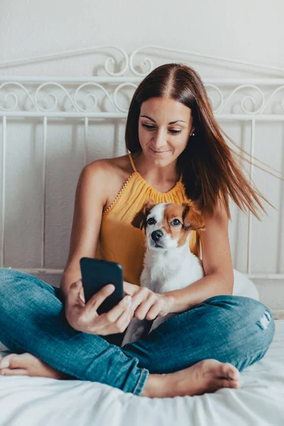 ผู้หญิงผิวขาวสวยนั่งบนเตียงกับสุนัขของเธอ แจ็ค รัสเซล ดูโทรศัพท์มือถือ — ภาพถ่ายสต็อก
