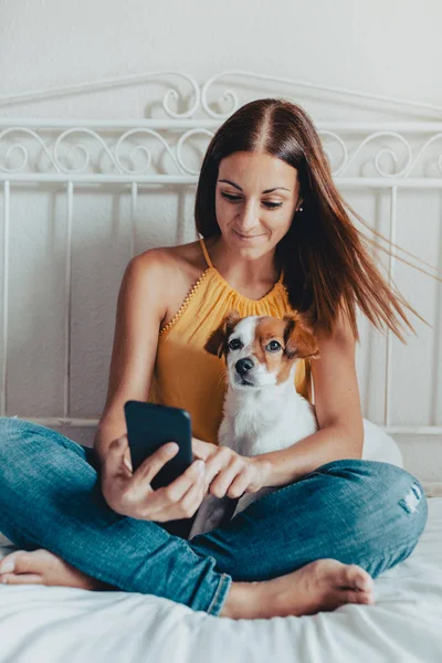 Belle femme caucasienne assise au lit avec son chien Jack Russell regardant le mobile Image En Vente