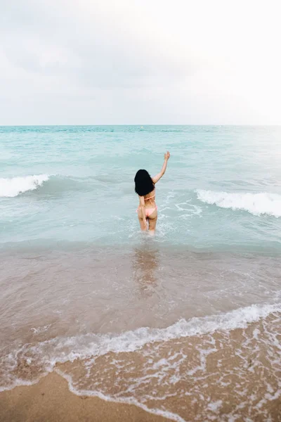 Vista posterior de la mujer sexy con bikini rosa jugando en la playa de agua cristalina azul en un día nublado. Concepto de viaje . — Foto de Stock