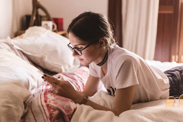 สาวสวยที่มีแว่นตานอนบนเตียงและมองหาบางอย่างบนโทรศัพท์มือถือ — ภาพถ่ายสต็อก