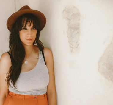 Beyaz duvara şapka takan genç esmer kadın kameraya bakıyor. Seyahat konsepti