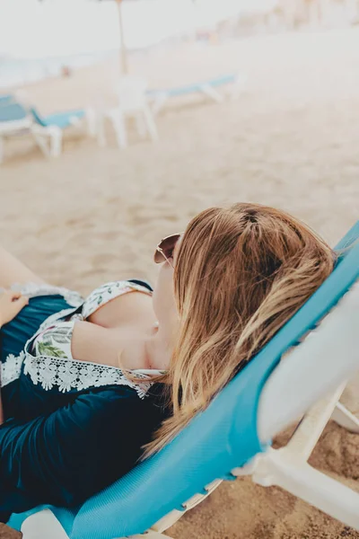 Giovane donna bionda sdraiata in un lettino blu con occhiali da sole sorridente godendo la spiaggia — Foto Stock