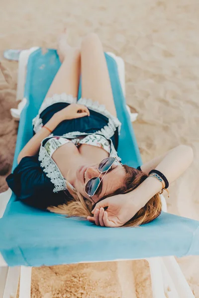 Giovane donna bionda con i capelli corti sdraiata su un'amaca blu con occhiali da sole guardando la fotocamera e godendo la spiaggia — Foto Stock
