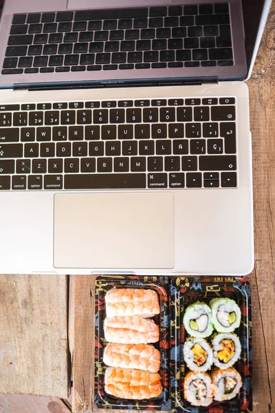 ม้วนซูชิอร่อยกุ้งและแล็ปท็อปที่โต๊ะไม้ จัดส่งอาหาร พื้นที่ทํางานที่บ้าน — ภาพถ่ายสต็อก