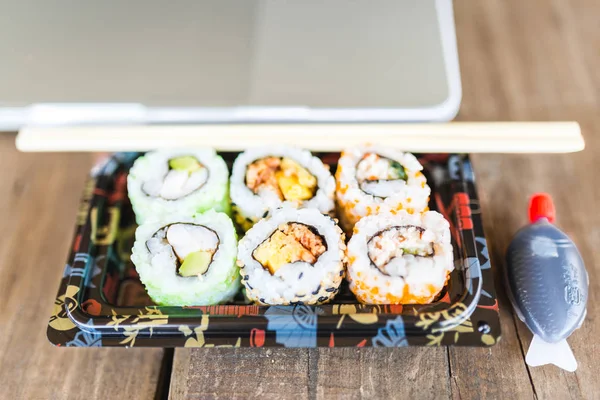ม้วนซูชิอร่อยและแล็ปท็อปที่โต๊ะไม้ จัดส่งอาหาร สถานที่ทํางานที่บ้าน — ภาพถ่ายสต็อก