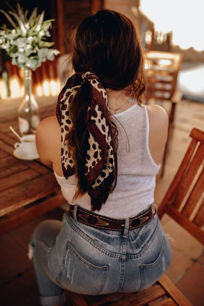 มุมมองด้านหลังของหญิงสาวสีน้ําตาลเซ็กซี่ที่มีหางม้าและ bandana นั่งอยู่บนหลังของเธอบนระเบียงดื่มกาแฟหนึ่งถ้วย . — ภาพถ่ายสต็อก