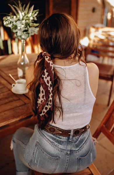 Вид сзади на сексуальную молодую женщину с хвостиком и повязкой на спине, сидящую на террасе и пьющую чашку кофе во второй половине дня . Стоковое Изображение