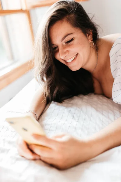 ผู้หญิงที่มีความสุขที่บ้านส่งข้อความในโทรศัพท์มือถือของเธอในห้องนอนที่สดใส ตรวจสอบแอพโซเชียลด้วยสมาร์ทโฟน คอนเซ็ปต์ไลฟ์สไตล์ . — ภาพถ่ายสต็อก