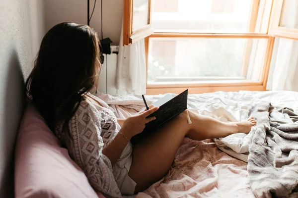 Side view of woman writing in a notebook on bed at home at evening. Close up lifestyle of pretty young woman sitting on her cozy bed and making notes to her diary. Fall season mood. Bright colors. — ストック写真