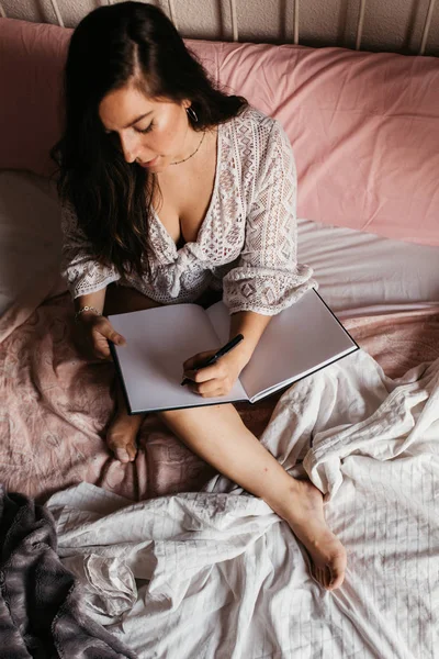 Top view of young brunette woman writing in a notebook on bed at — ストック写真