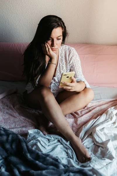Young sad woman sitting on the bed looking at her smartphone feels unhappy. Waiting for mobile message. Feeling worried, hurt, heartbroken lonely ignored by boyfriend not texting on cellphone — ストック写真