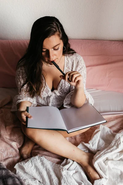 ท็อปวิวของหญิงสาวสีน้ําตาลคิดและเขียนในสมุดบันทึกบนเตียงที่บ้านในตอนเช้า ผู้หญิงเพลิดเพลินกับนิยายในห้องนอนแนวคิดการใช้ชีวิตการศึกษา ผู้หญิงสาวอ่านหนังสือบนเตียงที่บ้าน — ภาพถ่ายสต็อก