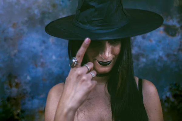 Een jonge gelukkige Halloween heks die glimlacht en haar heksenhoed aanraakt. Schoonheidsheks met zwarte mond in het donker. Serieus model meisje met kostuum en Halloween make-up. Donkere achtergrond. — Stockfoto