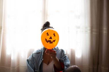 Cadı eliyle balkabağı tutuyor ve yüzünü örtüyor. Cadılar bayramını kutlayan genç bir kadın. Helyum parti balonları.