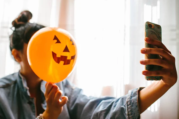 Bruxa segurando um balão de abóbora com a mão, cobrindo o rosto e tirando uma foto. Uma jovem que celebra o Halloween. Balões de festa de hélio . — Fotografia de Stock