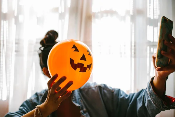 Bruxa segurando um balão de abóbora com a mão, cobrindo o rosto e tirando uma foto com celular. Uma jovem que celebra o Halloween. Balões de festa de hélio . — Fotografia de Stock