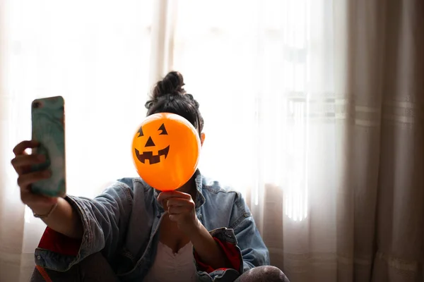 Bruxa segurando um balão de abóbora com a mão, cobrindo o rosto e tirando uma foto com celular. Uma jovem que celebra o Halloween. Balões de festa de hélio . — Fotografia de Stock